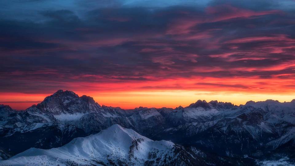 Jagged snow-covered mountain peaks under a dramatic sunset sky with orange, red, and purple clouds
