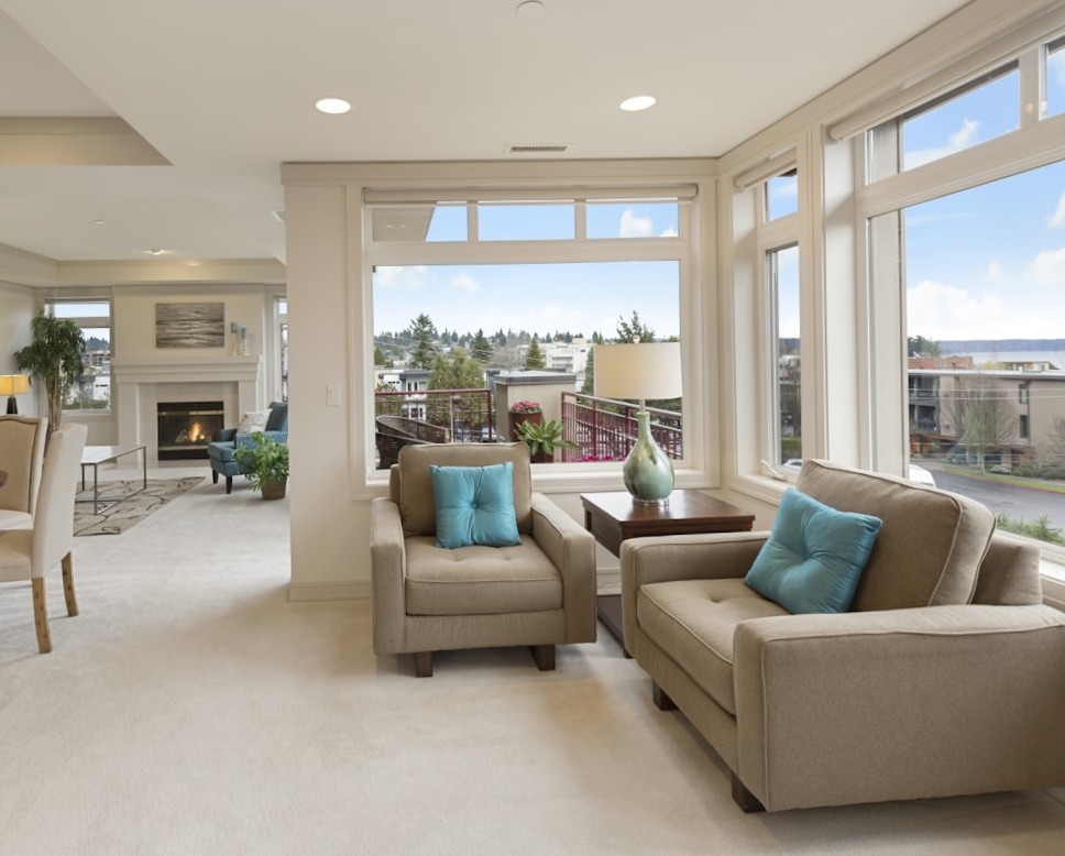 Modern living room with beige armchairs, turquoise pillows, and floor-to-ceiling windows facing a lake